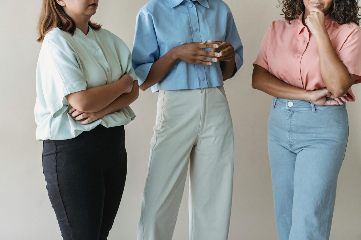 Three women in pastel shirts and pants stand together indoors, embodying casual, pastel fashion.